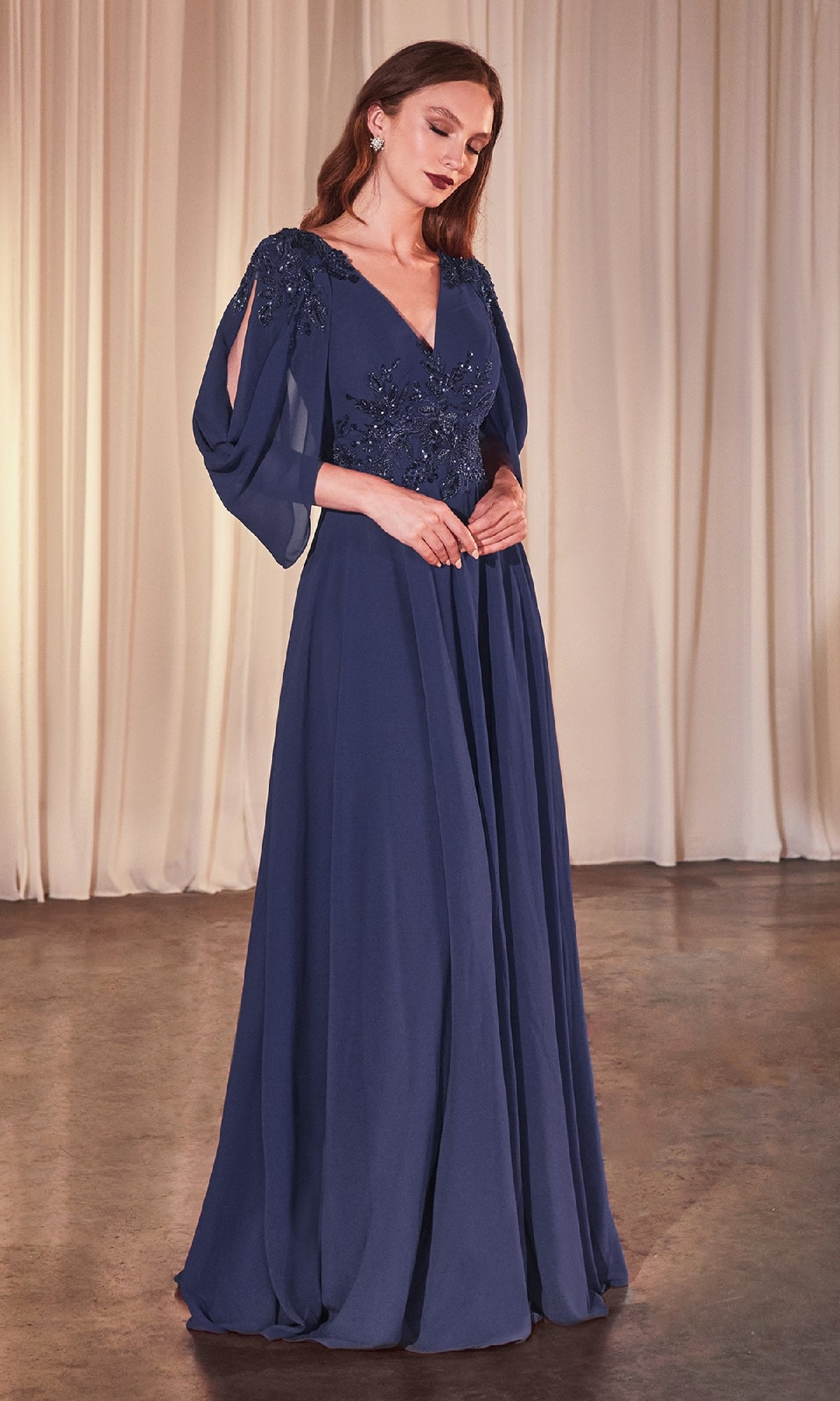 A woman stands in front of cream curtains wearing the Ladivine CDS508, a floor-length navy blue formal dress featuring beaded bodice detailing and elegant cowl sleeves.